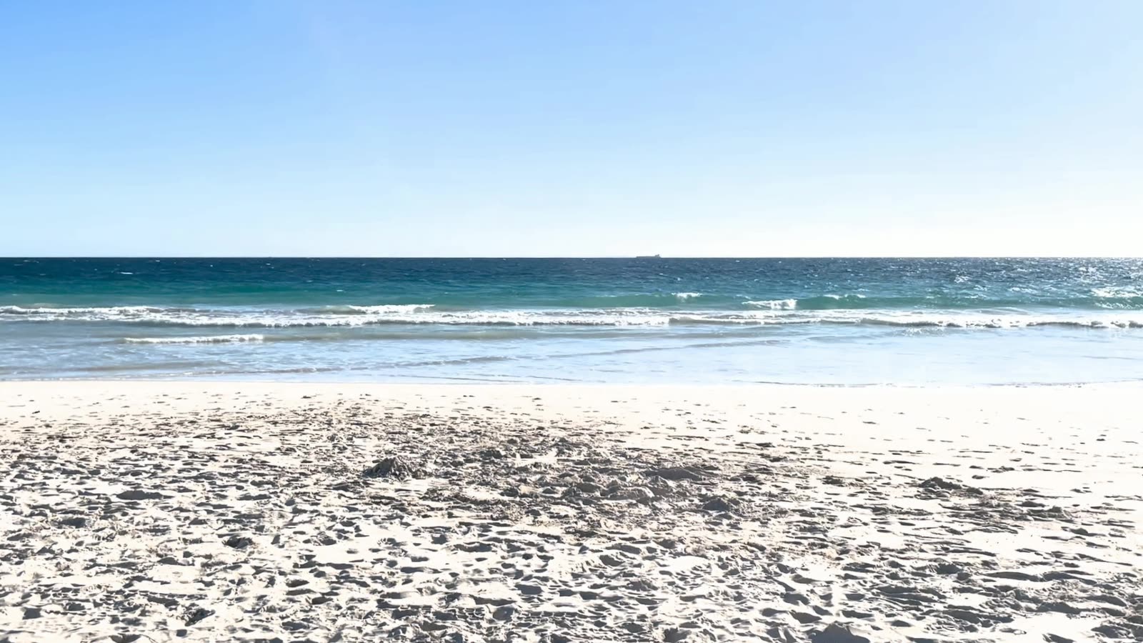 Waves breaking on a white sand beach, filmed from a seated position, with a soft light half set on the horizon